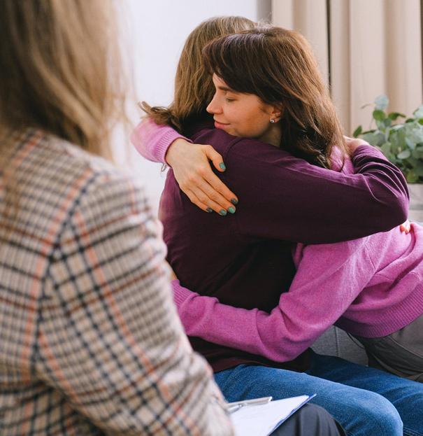 Two individuals embracing during a counseling session, with a therapist observing in the background, illustrating how Debbie Bayer LMFT provides compassionate and supportive relationship counseling services.